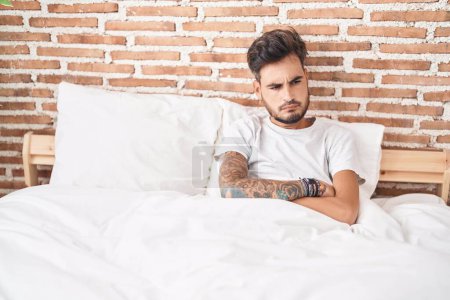 Foto de Young hispanic man sitting on bed with unhappy expression and arms crossed gesture at bedroom - Imagen libre de derechos