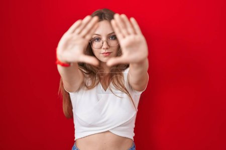 Photo for Young caucasian woman standing over red background doing frame using hands palms and fingers, camera perspective - Royalty Free Image