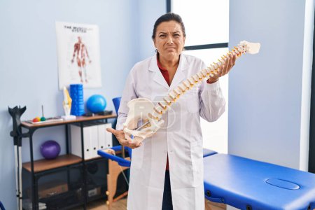 Foto de Mujer hispana de mediana edad que sostiene el modelo anatómico de la columna vertebral expresión despistada y confusa. concepto de duda. - Imagen libre de derechos