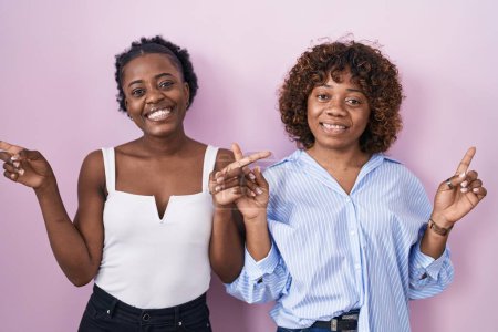 Téléchargez les photos : Deux femmes africaines debout sur fond rose souriant confiant pointant avec les doigts vers des directions différentes. espace de copie pour la publicité - en image libre de droit