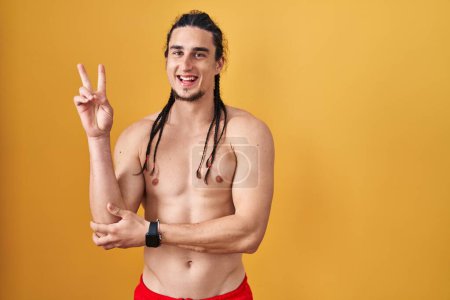 Téléchargez les photos : Homme hispanique avec les cheveux longs debout torse nu sur fond jaune souriant avec le visage heureux clin d'oeil à la caméra faisant signe de victoire avec les doigts. numéro deux. - en image libre de droit
