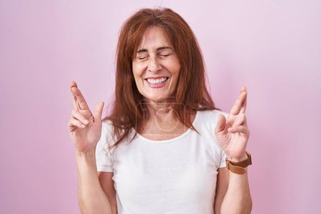 Téléchargez les photos : Middle age woman standing over pink background gesturing finger crossed smiling with hope and eyes closed. luck and superstitious concept. - en image libre de droit