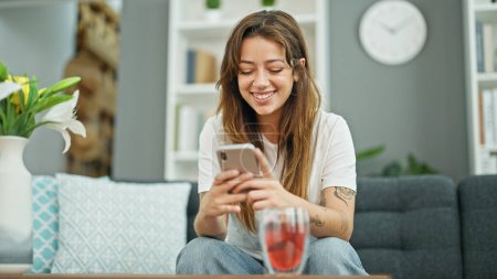 Foto de Mujer hispana hermosa joven usando teléfono inteligente sentado en el sofá sonriendo en casa - Imagen libre de derechos