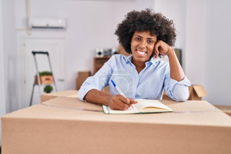 Téléchargez les photos : African american woman smiling confident writing on notebook at new home - en image libre de droit