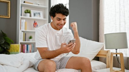 Téléchargez les photos : Jeune homme latino utilisant smartphone assis sur le lit célébrant dans la chambre - en image libre de droit