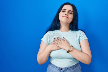 Photo for Young modern girl with blue hair standing over blue background smiling with hands on chest with closed eyes and grateful gesture on face. health concept. - Royalty Free Image