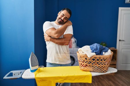 Foto de Joven hispano con barba planchando ropa en casa abrazándose feliz y positivo, sonriendo confiado. amor propio y cuidado personal - Imagen libre de derechos