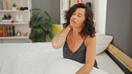 Foto de Mujer hispana de mediana edad sentada en la cama sufriendo de dolor cervical en el dormitorio - Imagen libre de derechos