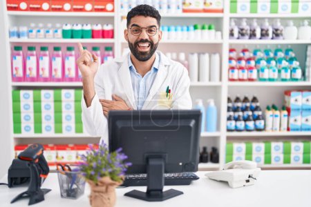 Photo for Hispanic man with beard working at pharmacy drugstore with a big smile on face, pointing with hand and finger to the side looking at the camera. - Royalty Free Image