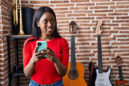 Foto de Joven afroamericana mujer músico sonriendo confiado usando smartphone en estudio de música - Imagen libre de derechos