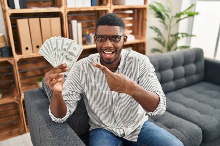 Téléchargez les photos : Africain américain tenant des dollars souriant heureux pointant avec la main et le doigt - en image libre de droit