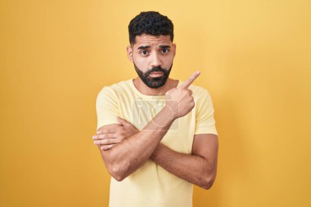 Foto de Hombre hispano con barba de pie sobre fondo amarillo señalando con el dedo de la mano a un lado mostrando publicidad, cara seria y tranquila - Imagen libre de derechos