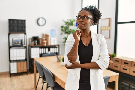 Téléchargez les photos : Femme afro-américaine ouvrière d'entreprise debout avec l'expression du doute au bureau - en image libre de droit