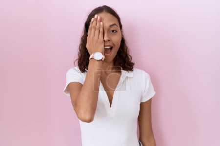 Téléchargez les photos : Jeune femme hispanique portant t-shirt blanc décontracté couvrant un oeil avec la main, sourire confiant sur le visage et l'émotion surprise. - en image libre de droit