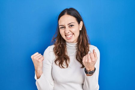 Foto de Young hispanic woman standing over blue background very happy and excited doing winner gesture with arms raised, smiling and screaming for success. celebration concept. - Imagen libre de derechos