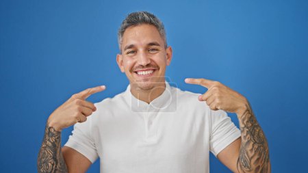 Foto de Joven hombre hispano sonriendo confiado señalando a la boca sobre aislado fondo azul - Imagen libre de derechos