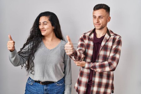 Photo for Young hispanic couple standing over white background looking proud, smiling doing thumbs up gesture to the side - Royalty Free Image