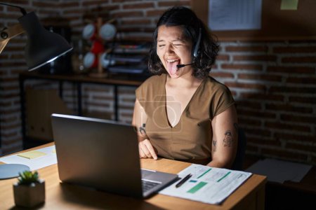 Photo for Young hispanic woman working at the office at night sticking tongue out happy with funny expression. emotion concept. - Royalty Free Image