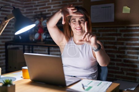 Téléchargez les photos : Femme brune travaillant au bureau la nuit souriant faisant cadre avec les mains et les doigts avec le visage heureux. concept de créativité et photographie. - en image libre de droit
