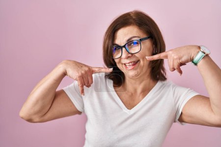 Téléchargez les photos : Middle age hispanic woman standing over pink background smiling cheerful showing and pointing with fingers teeth and mouth. dental health concept. - en image libre de droit