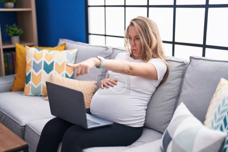 Téléchargez les photos : Jeune femme enceinte assise sur le canapé à la maison à l'aide d'un ordinateur portable pointant du doigt la caméra et à vous, geste confiant regardant sérieux - en image libre de droit