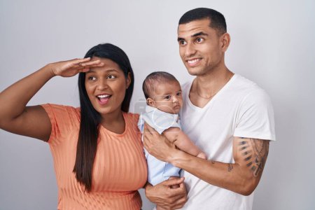 Photo for Young hispanic couple with baby standing together over isolated background very happy and smiling looking far away with hand over head. searching concept. - Royalty Free Image