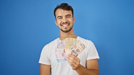 Foto de Joven hombre hispano sonriendo confiado sosteniendo pesos mexicanos sobre aislado fondo azul - Imagen libre de derechos