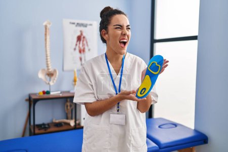 Foto de Mujer hispana joven sosteniendo plantilla de zapato en clínica de fisioterapia enojada y loca gritando frustrada y furiosa, gritando de rabia. rabia y concepto agresivo. - Imagen libre de derechos
