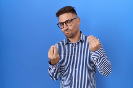 Foto de Hombre hispano con barba usando gafas haciendo gesto de dinero con las manos, pidiendo pago de sueldo, negocio millonario - Imagen libre de derechos