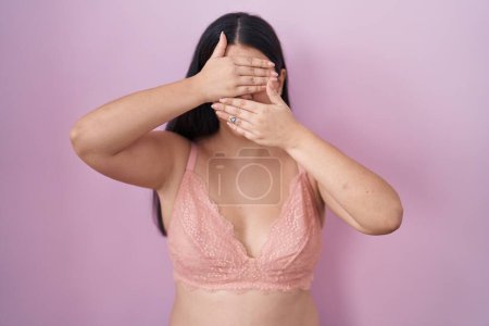 Foto de Mujer hispana joven con sujetador rosa cubriendo los ojos y la boca con las manos, sorprendida y conmocionada. ocultar emociones - Imagen libre de derechos