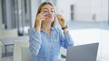 Téléchargez les photos : Jeune femme blonde femme d'affaires avec stylo entre le nez et la bouche faisant un geste drôle au bureau - en image libre de droit