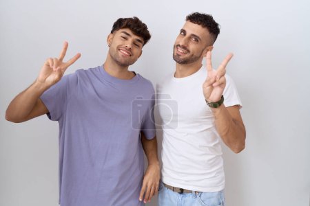 Téléchargez les photos : Couple homosexuel gay debout sur fond blanc souriant en regardant la caméra montrant les doigts faisant signe de victoire. numéro deux. - en image libre de droit