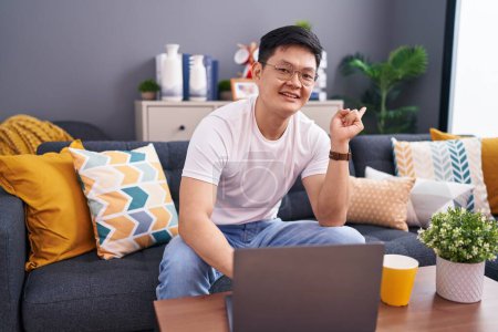 Téléchargez les photos : Jeune homme asiatique à l'aide d'un ordinateur portable à la maison assis sur le canapé avec un grand sourire sur le visage, pointant avec le doigt de la main sur le côté regardant la caméra. - en image libre de droit