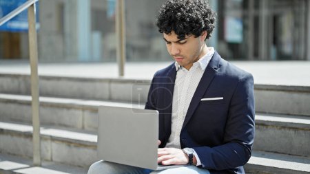 Téléchargez les photos : Jeune homme d'affaires latino-américain utilisant un ordinateur portable assis sur le sol de la rue - en image libre de droit