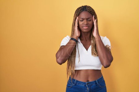 Téléchargez les photos : Femme afro-américaine avec les cheveux tressés debout sur fond jaune avec la main sur la tête, maux de tête parce que le stress. migraine souffrant. - en image libre de droit