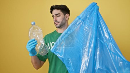 Téléchargez les photos : Jeune homme hispanique volontaire portant des gants recyclage bouteille en plastique sur fond jaune isolé - en image libre de droit