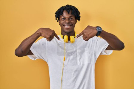 Téléchargez les photos : Jeune homme africain avec dreadlocks debout sur fond jaune regardant confiant avec le sourire sur le visage, se montrant du doigt fier et heureux. - en image libre de droit