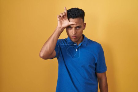 Photo for Young hispanic man standing over yellow background making fun of people with fingers on forehead doing loser gesture mocking and insulting. - Royalty Free Image