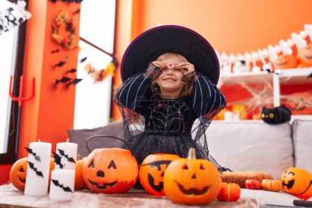 Foto de Adorable chica rubia con disfraz de bruja sosteniendo dulces sobre los ojos en casa - Imagen libre de derechos