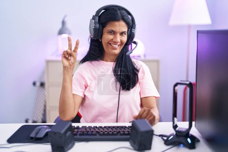Foto de Mujer hispana madura jugando videojuegos en casa mostrando y señalando con los dedos número dos mientras sonríe confiada y feliz. - Imagen libre de derechos