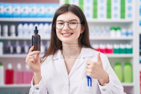 Téléchargez les photos : Jeune femme caucasienne travaillant à la pharmacie drugstore tenant sérum souriant heureux et positif, pouce levé faire excellent et signe d'approbation - en image libre de droit