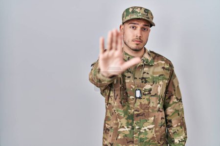 Foto de Joven hispano con uniforme camuflaje del ejército haciendo parar de cantar con la palma de la mano. expresión de advertencia con gesto negativo y grave en la cara. - Imagen libre de derechos