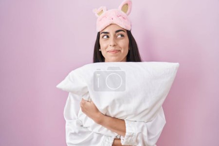 Téléchargez les photos : Jeune femme brune portant un masque de sommeil et un pyjama embrassant un oreiller souriant regardant vers le côté et regardant loin en pensant. - en image libre de droit