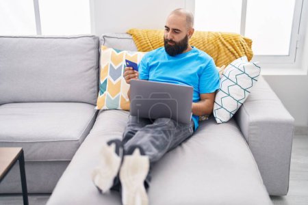Téléchargez les photos : Jeune chauve utilisant ordinateur portable et carte de crédit assis sur le canapé à la maison - en image libre de droit