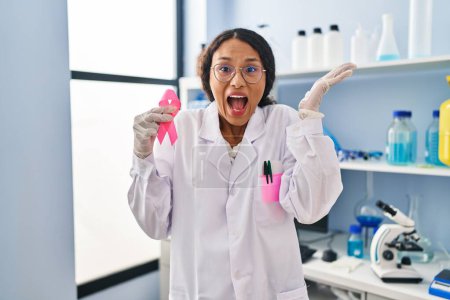 Photo for Young hispanic doctor woman working at scientist laboratory holding pink ribbon celebrating victory with happy smile and winner expression with raised hands - Royalty Free Image