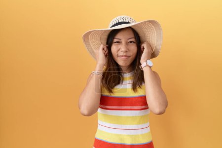 Foto de Mujer china de mediana edad con sombrero de verano sobre fondo amarillo que cubre las orejas con los dedos con expresión molesta por el ruido de la música fuerte. concepto de sordo. - Imagen libre de derechos