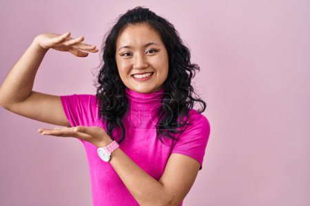 Foto de Joven mujer asiática de pie sobre fondo rosa gesto con las manos mostrando signo de tamaño grande y grande, símbolo de medida. sonriendo mirando a la cámara. concepto de medición. - Imagen libre de derechos