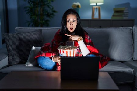 Téléchargez les photos : Femme hispanique manger pop-corn regarder un film sur le canapé surpris pointant du doigt sur le côté, bouche ouverte expression étonnante. - en image libre de droit