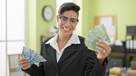 Foto de Joven hermosa mujer hispana trabajadora de negocios sosteniendo pesos chilenos sonriendo en la oficina - Imagen libre de derechos