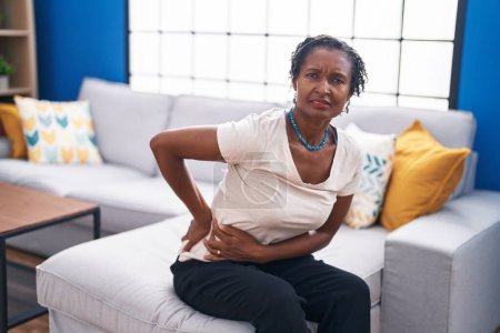 Téléchargez les photos : Moyen Age femme afro-américaine souffrant de maux de dos assis sur le canapé à la maison - en image libre de droit